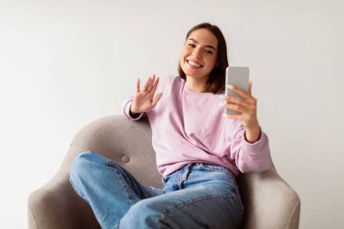 Sosyal ağlar, görüntülü konuşma, blog yazma, canlı yayın. Sırıtkan beyaz kadın koltukta akıllı telefon kullanıyor, webcam 'e el sallıyor, beyaz arka planda arkadaşlarıyla veya aboneleriyle konuşuyor.