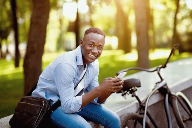 Şehir parkında bisikletli mutlu siyah adam akıllı telefondan konuşuyor.