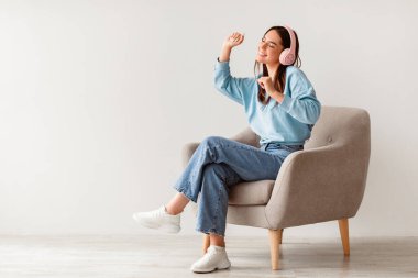 Kablosuz kulaklıklı, koltukta dinlenen ve beyaz stüdyo duvarına karşı müziğin keyfini çıkaran mutlu bir milenyum kadını. Tatlı genç kadın en sevdiği şarkı listesiyle dans ediyor