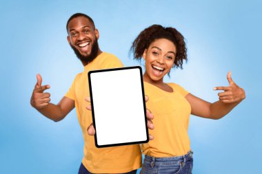 Neşeli Afrikalı Amerikalı çift mavi stüdyo arka planında tasarım, yeni uygulama veya web sitesi için tablet ekranını işaret ediyor. İlanınız için dokunun ekran şablonu, boşluk