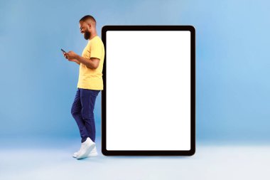 Beyaz ekranlı dev dijital tabletin yanında duran çekici Afrikalı Amerikalı adam, akıllı telefon kullanıyor, mavi stüdyo arka planında yeni uygulamalar tanıtıyor, web sitesi veya reklam tasarımı için modelleme yapıyor.