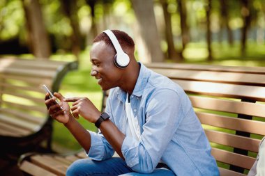 Parktaki bankta müzik listesi seçen kulaklıklı çekici Afrikalı Amerikalı adam.