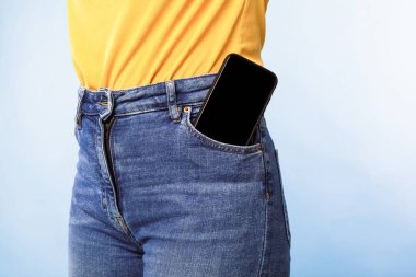 Mavi stüdyo arka planında pantolonunun cebinde boş ekranı olan akıllı telefonu olan tanınmayan bir kadın. Mobil uygulama ya da web sitesi tasarımı için model. İlan şablonunuz için boşluk