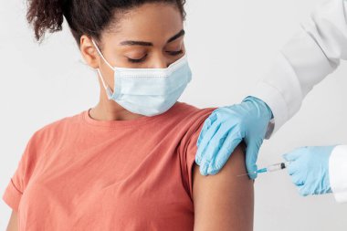 Beyaz iş elbiseli hemşire, eldivenler ve koruyucu yüz maskesi hastanede, klinikte veya aşı merkezinde genç siyahi bir kadın için omuzlarında koronavirüs aşısı yapıyor. Güvenlik bağışıklığı kavramı