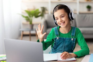 Mesafe öğrenme. Kulaklık takmış, video dersi alan, bilgisayarını oturma odasında kullanan, uzaktan eğitim alan, online eğitimin tadını çıkaran, bilgisayar kamerasına el sallayan sevimli bir genç kız.