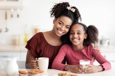 Genç siyah anne ve genç kızın taze ev yapımı kurabiyelerin tadını çıkarması, kahve ve süt içmesi, mutfakta tabletin başında oturup kameraya gülümsemesi, evde birlikte vakit geçirmesi, yer kopyalaması.