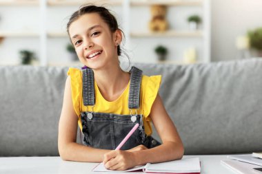 Modern Eğitim Konsepti. Oturma odasında oturan gülümseyen küçük kız öğrencinin portresi, defterine veya günlüğüne notlar yazması, kameraya poz vermesi, seçici odaklanma.