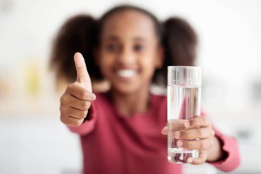 Seçici bir şekilde bir bardak suya odaklan ve başparmağını kaldır, tanınmayan siyah kız içme suyu iç, Afrikalı Amerikalı genç çocuk taze kaynak suyu tavsiye ediyor, yakın plan. Çocuklar için hidrasyon ilacı.