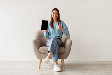 Gülümseyen milenyum kadını boş dokunmatik pedi gösteriyor, baş parmağını kaldırıyor, beyaz duvara yaslanmış koltukta oturuyor, tablet ekranda web sitesi tasarımı için maket yapıyor. Çevrimiçi reklam kavramı