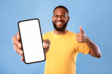Yeni havalı mobil uygulama. Funky Afrikalı Amerikalı adam cep telefonunu boş ekranla gösteriyor, başparmak hareketi gösteriyor, web sitesi tavsiye ediyor, mavi arka planda reklam için yer öneriyor,