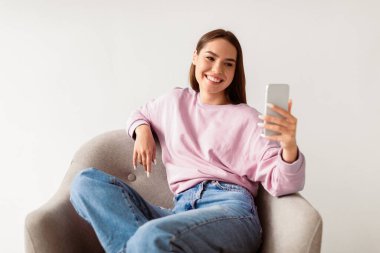 Akıllı telefon kullanan neşeli beyaz kadın internette sohbet ediyor, çalışıyor ya da internetten ders çalışıyor, beyaz stüdyo geçmişine karşı koltukta oturuyor. İnternet üzerinden iletişim kuran mobil cihazlı arkadaş canlısı bayan.