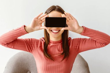 İnsanlar ve aletler. Beyaz stüdyo arka planında gözlerini cep telefonuyla kapayan neşeli genç bir bayan. Uygulama ya da cihaz ekranında web sitesi için bir model. Milenyum kadını dokunmatik ped reklamları için yer açıyor.