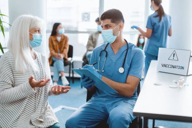 Doktor randevusu. Kıdemli hasta bayanla grip belirtileri hakkında konuşan adam, hastanede otururken yüz maskeleri takan, yan etkiler ve faydaları hakkında bilgi veren adam.