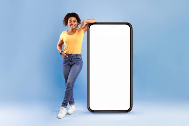Mobil uygulama reklamı. Beyaz ekranı boş, yeni web sitesi ya da uygulaması olan, mavi stüdyo geçmişi olan, büyük akıllı telefona yaslanan mutlu Afrikalı Amerikalı kadın.