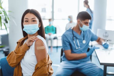 Aşı kavramından sonra. Aşılanmış, maskeli, koluna antiviral enjeksiyon yaptıran, modern klinikte kollarını sıvayıp kameraya bakan genç bir kadının portresi.