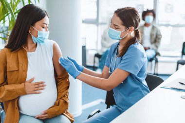 Yüz maskeli hemşire ve hamile bir hastanın derisini dezenfekte eden eldivenler, enjeksiyondan önce omuzlarını pamukla ovmak, Asyalı bir kadının göbeğine sarılması, klinik geçmişi. Grip koronavirüsü tedavisi kavramı