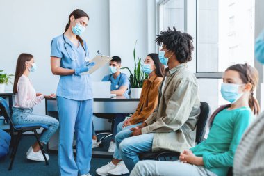 Korumacı yüz maskesi takmış insanlar, çoklu aşıya karşı aşı bekliyorlar, kuyrukta bekliyorlar, çok ırklı hastalar klinikte aşılanmaya hazırlanıyorlar, hemşire yazıları yazıyorlar.