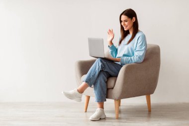 Beyaz stüdyo duvarına karşı koltukta online iletişim için dizüstü bilgisayar kullanan beyaz kadının tam boy portresi, kopyalama alanı. Güzel genç bir kadın arkadaşıyla görüntülü konuşma yapıyor, web kamerasına el sallıyor.