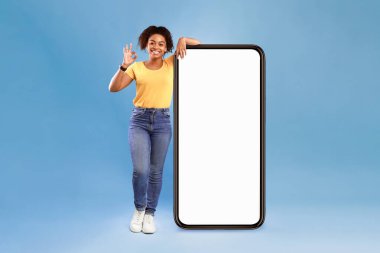 Yeni harika mobil uygulama. Büyük bir cep telefonunun yanında boş ekranla poz veren Afro-Amerikalı bir kadın, iyi bir jest gösteriyor, web sitesi tavsiye ediyor, reklamlar için mavi ekranda yer açıyor.