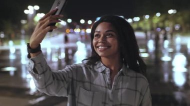 Genç bir Afro-Amerikalı kadın gece canlı bir sokakta tek başına yürüyor, selfie çekerken gülümsüyor. Şehir ışıkları ıslak asfalttan yansıyor, atmosfer rahat..