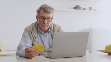 Parlak ve modern bir yaşam alanında, gri saçlı yaşlı bir adam bir masada oturuyor, elinde bir kredi kartı tutarken dizüstü bilgisayarına odaklanıyor, dijital işlemlerden keyif alıyor..