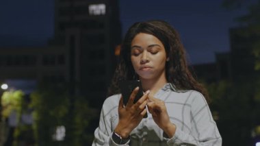 Genç bir Afrikalı Amerikalı kadın gece loş bir sokakta akıllı telefonuna odaklanarak geziniyor. Akşam havası serin, yalnızlığının ve çevrenin tadını çıkarıyor..