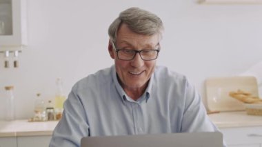 60 'lı yaşlarda yaşlı bir adam mutfak masasında dizüstü bilgisayarına odaklanmış oturuyor. Gözlük takıyor ve rahat giyiniyor. Etrafı düzenli bir ev ortamıyla çevrili. İfadesi konsantrasyonu gösteriyor..