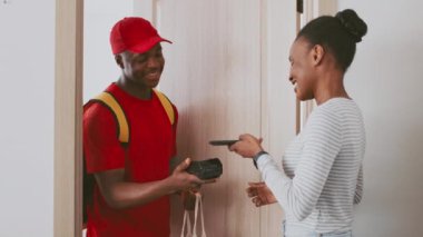 Kırmızı gömlekli ve şapkalı bir adam dairesindeki gülümseyen bir kadına paket teslim ediyor. Arkadaşça etkileşim kurye hizmetleri ve müşteriler arasındaki bağlantıyı vurgular.