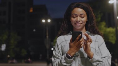 Genç bir Afro-Amerikalı kadın, gece sokakta akıllı telefonunu tutarak yürüyor ve gülümsüyor. Mesajlarını dinlerken ve müzik dinlerken huzurlu bir akşamın tadını çıkarıyor..