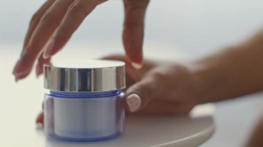 Genç bir Afrikalı Amerikalı kadın evdeki şımartma rutini sırasında elini nazikçe bir kavanoz cilt kremine koyuyor ve bir anlık özgüven ve rahatlama anının tadını çıkarıyor..