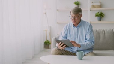 60 'lı yaşlarda yaşlı bir adam kanepede rahatça oturur ve tabletine göz atar. Konforlu ev ortamında hoş bir dekor ve yeşillik var. Huzurlu bir atmosfer yaratıyor..