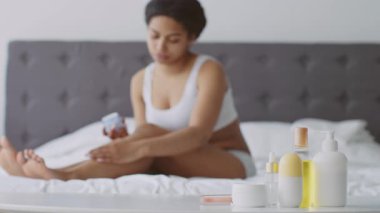 Genç bir Afrikalı Amerikalı kadın, evdeki rahatlatıcı güzellik rutininin bir parçası olarak rahatça yatağında oturup cilt bakım ürünleri uyguluyor. Sakin bir ortamda kendine bakmanın tadını çıkarıyor..