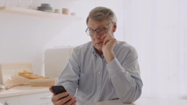 60 'lı yaşlarda parlak bir mutfakta oturan, akıllı telefonuna odaklanmış son sınıf öğrencisi. Gri saçları ve gözlükleri var. Huzurlu bir ortamda geçirdiği emeklilik yıllarını yansıtıyor..