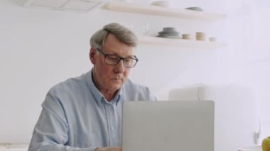 60 'lı yaşlarda emekli bir adam, gözlük takıyor, parlak bir mutfakta dizüstü bilgisayarına odaklanıyor. O, evdeki emeklilik yaşam tarzının basitliğinin tadını çıkarmakla meşgul..