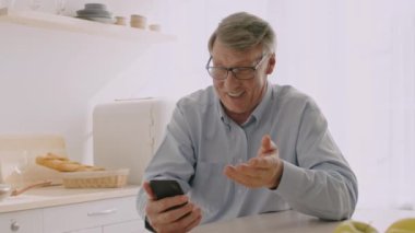 60 'lı yaşlarında yaşlı bir adam mutfak masasında akıllı telefonuyla meşgul olarak oturuyor. Gri saçı, rahat ve aydınlık bir ev ortamında rahat tavırlarını tamamlıyor..