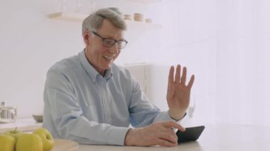 60 'lı yaşlarda yaşlı gri saçlı bir adam evdeki bir video görüşmesi sırasında gülümsüyor ve el sallıyor. Elmalarla çevrili bir mutfak masasında rahatça oturuyor ve neşeli bir çevresi var..