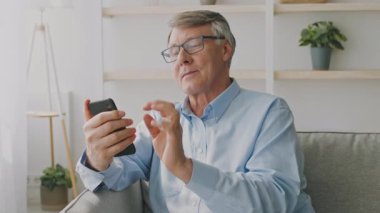 60 'larında yaşlı bir adam rahat bir oturma odasında rahat bir koltukta oturuyor, akıllı telefonuyla meşgul. Uygulamalarda gezinirken mutlu görünüyor, emekliliğinin tadını çıkarıyor..