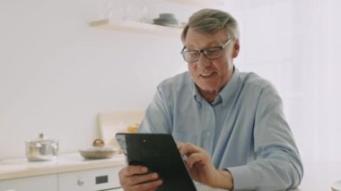 Bir tabletle meşgul olurken gri saçlı, 60 'lı yaşlarda emekli bir adam gülümsüyor. Parlak, rahat bir mutfakta oturmuş, rahat bir atmosferde eğleniyor..