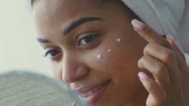 Bu genç Afrikalı Amerikalı kadın cildine krem sürüyor. Parlak ve huzurlu bir evde. Kendini pohpohlayan, kendine güveni ve güzelliği yayarak cildine özen gösterir..