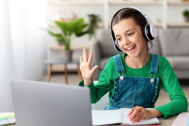 Çevrimiçi Görüntülü Arama ve İletişim. Masada oturmuş bilgisayarla sanal konferans yapan neşeli ergen öğrencinin portresi. Web kameralı selamlama öğretmenine ve sınıf arkadaşlarına el sallıyor.