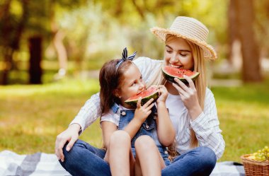 Küçük kız ve annesi, Sümer pikniğinde karpuz yiyorlar. Parkta oturmuş, hafta sonunu birlikte geçiriyorlar..