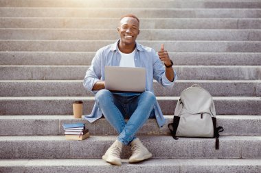 Laptoplu pozitif siyah adam şehir merkezindeki merdivenlerde baş parmak hareketi gösteriyor.