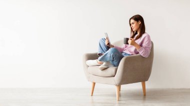 Koltuğunda oturan, beyaz stüdyo duvarına karşı tablet bilgisayar kullanan, kahvesi boş bir panorama olan beyaz bir kadın. Milenyum bayanı dokunmatik ped üzerinde çalışıyor ya da okuyor