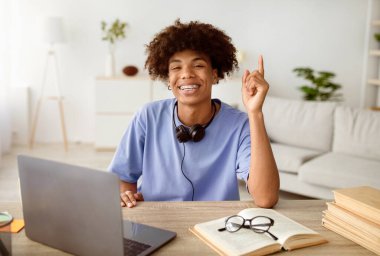 Kulaklık takan, dizüstü bilgisayarının yanında yaratıcı fikirleri olan mutlu siyahi öğrenci evde Eureka 'yı işaret ediyor. Afro ergen çevrimiçi eğitim kursu için ödev yaparken...