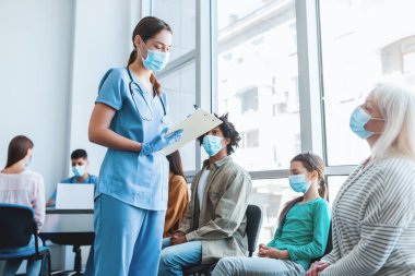 Çeşitli insanlar sırada oturup hastane aşılama merkezinde sıralarının gelmesini beklerler. Hemşire antiviral grip aşısı kuyruğundaki hastalarla konuşuyor.