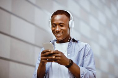 Neşeli Afro-Amerikan kulaklıklı adam dışarıdaki duvarın yanındaki akıllı telefondan müzik listesi seçiyor.