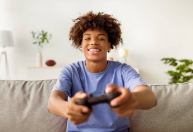 Ev içi eğlenceler. Evdeki kanepede elinde joystick ile bilgisayar oyunu oynayan neşeli siyahi bir genç. Video oyunlarıyla uğraşan Afrikalı Amerikalı bir ergen.