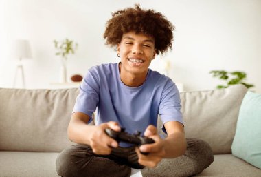 Evde eğleniyorum. Oyun kumandalı neşeli siyahi genç adam çevrimiçi bilgisayar oyunları oynuyor, içeride kanepede oturuyor. Coronavirus karantinası sırasında kontrol amaçlı video oyunu oynayan Afro gençliğinin portresi