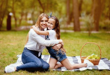 Mutlu anne ve kız ekose üzerinde oturup parkta piknik yapıyor, kameraya gülümsüyor. Yaz Haftasonu. Boş Boşluk