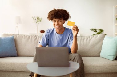 Uzaktan alışveriş konsepti. Bilgisayarı ve kredi kartıyla evden mal sipariş eden pozitif siyah bir genç. Neşeli Afrikalı Amerikalı ergen oturma odasında internetten bir şeyler alıyor.